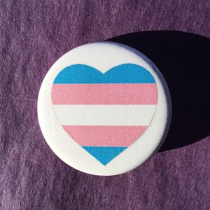 May include: A white button with a transgender pride flag in the shape of a heart. The flag is blue, pink, white, pink, and blue.