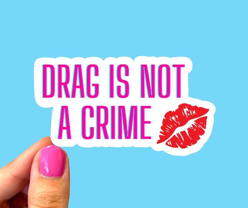 May include: White sticker with pink text that reads "DRAG IS NOT A CRIME" and a red lip print.