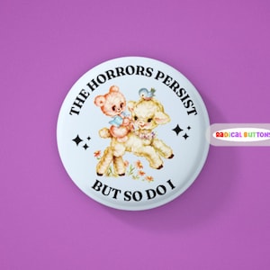 May include: A white button with the text "THE HORRORS PERSIST BUT SO DO I" in a circular design. The button features a vintage illustration of a lamb and a teddy bear. The button also has the text "RADICAL BUTTONS".