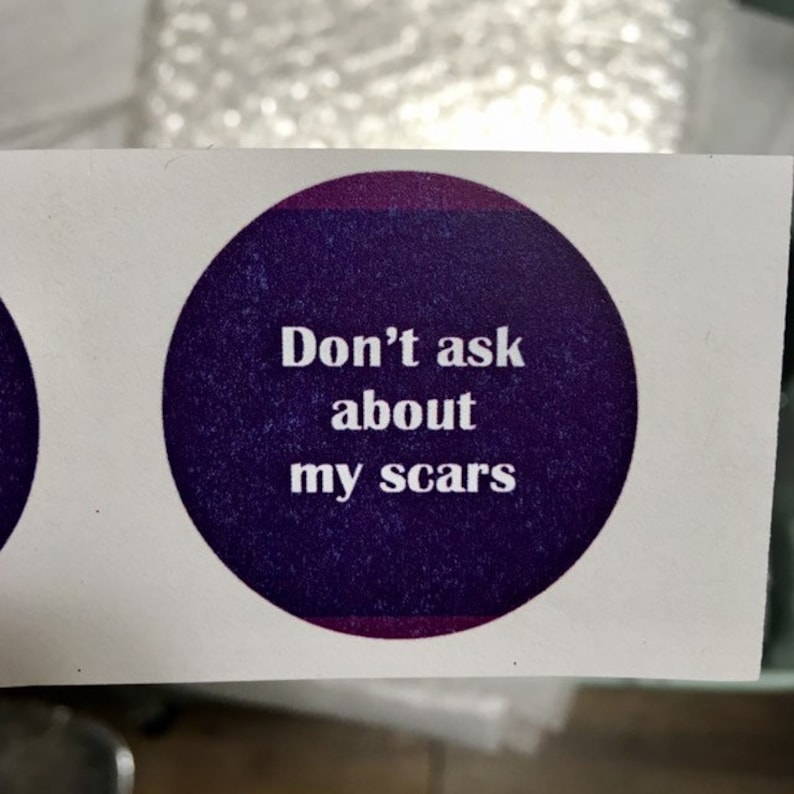 Puede incluir: Una pegatina redonda con el texto "Don't ask about my scars" en fuente blanca. La pegatina tiene un fondo morado oscuro con un borde morado m&aacute;s claro y est&aacute; sobre un fondo blanco.