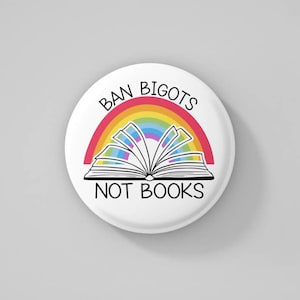 May include: A round white button featuring the text "BAN BIGOTS NOT BOOKS" in black. A rainbow design arches over an open book illustration. The button has a glossy finish.
