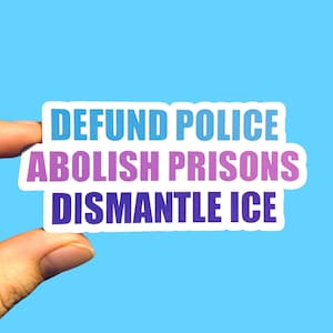 May include: A white sticker with the words "DEFUND POLICE, ABOLISH PRISONS, DISMANTLE ICE" in blue, purple, and dark blue text. The sticker is held between two fingers against a light blue background.
