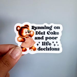 May include: A die-cut sticker featuring a brown teddy bear wearing a red and white checkered dress with a small butterfly on its nose. The text reads Running on Diet Coke and poor life decisions, accompanied by four small black star icons.