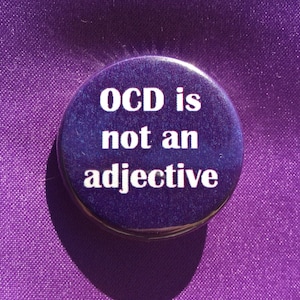 Puede incluir: Un bot&oacute;n redondo, de color p&uacute;rpura oscuro, con el texto blanco "OCD is not an adjective". El bot&oacute;n est&aacute; sobre un fondo de tela p&uacute;rpura. El bot&oacute;n tiene una superficie brillante y reflectante.