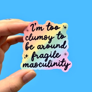 May include: A rainbow-colored sticker with the text "I'm too clumsy to be around fragile masculinity" in black lettering. The sticker has small star-shaped accents.