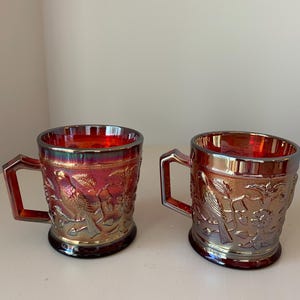 May include: Two iridescent red glass mugs with handles. The mugs feature embossed bird and floral designs. The glass has a shiny, metallic finish with hints of gold and purple. The mugs are likely vintage or antique.