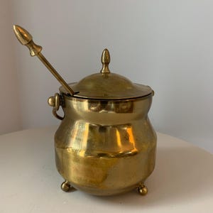 May include: A vintage brass sugar bowl with a lid and a small spoon. The bowl has a rounded body, three small feet, and a handle. The spoon rests inside the bowl. The brass has a warm, golden color.