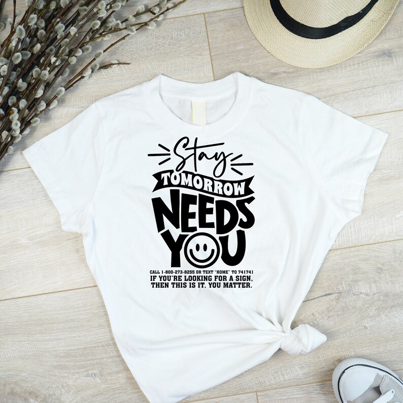 May include: White t-shirt with black text that reads "Stay Tomorrow Needs You" with a smiley face. The text also includes a phone number and the message "If you're looking for a sign, then this is it. You matter."