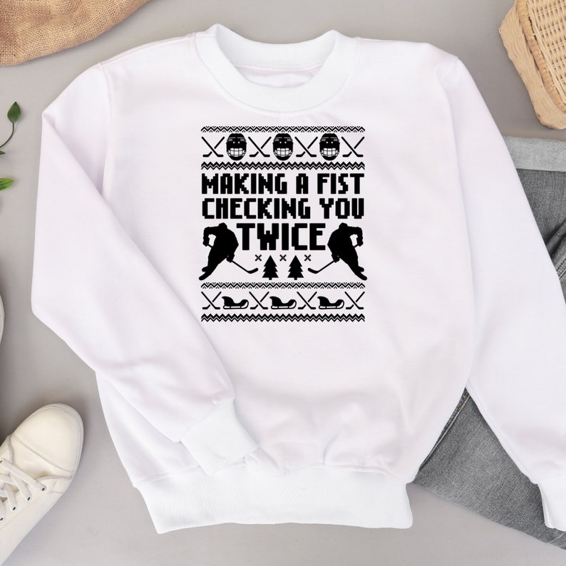 Puede incluir: Sudadera blanca con un dise&ntilde;o de hockey en blanco y negro. El dise&ntilde;o presenta dos jugadores de hockey, palos de hockey y el texto "Making a fist checking you twice".