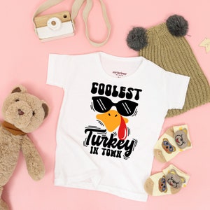May include: White baby t-shirt with a black and yellow turkey wearing sunglasses and the text "Coolest Turkey in Town".