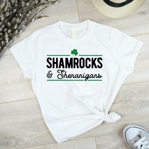 Op de afbeelding: Wit T-shirt met een groene klaver en de tekst "Shamrocks & Shenanigans" in zwart.