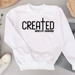 以下が含まれることがあります： 黒文字で「CREATED WITH A PURPOSE」と十字架のグラフィックが書かれた白いスウェットシャツです。