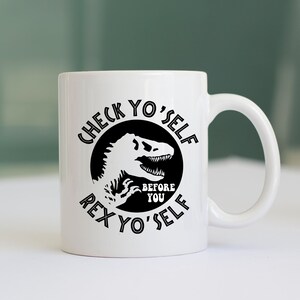 May include: White ceramic coffee mug with a black graphic of a dinosaur with the text "Check Yo'Self Before You Rex Yo'Self"