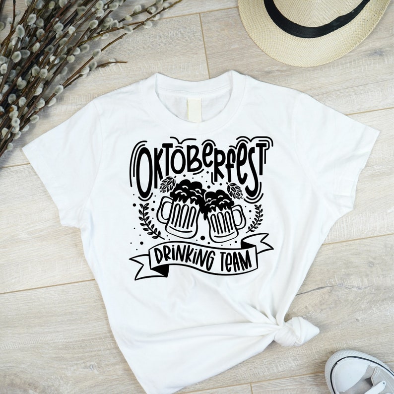 May include: White t-shirt with black text that says "Oktoberfest Drinking Team" and a graphic of two beer mugs with hops.