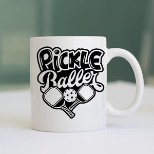 Pode incluir: Caneca de cer&acirc;mica branca com um design preto que diz "Pickle Baller" com duas raquetes de pickleball e uma bola.
