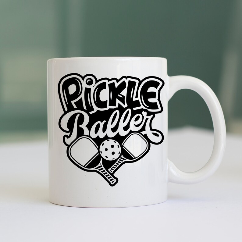 Pode incluir: Caneca de cer&acirc;mica branca com um design preto que diz "Pickle Baller" com duas raquetes de pickleball e uma bola.