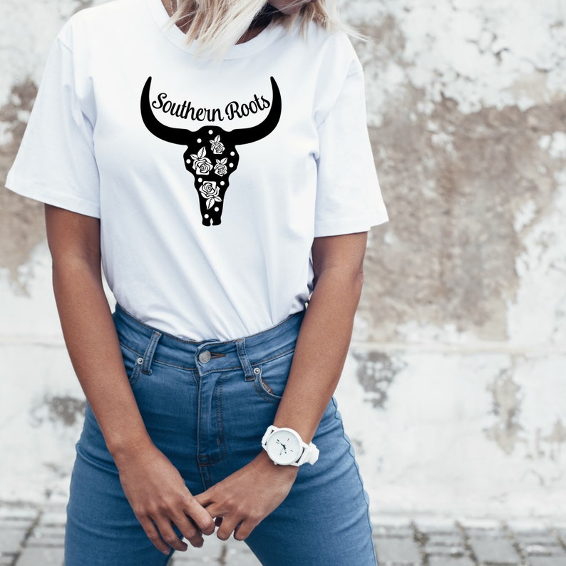 Puede incluir: Camiseta blanca con un dise&ntilde;o de cr&aacute;neo de toro negro y el texto "Southern Roots" impreso en la parte delantera.