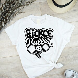 Pode incluir: Camiseta branca com texto preto que diz "Pickle Baller" e um gr&aacute;fico de duas raquetes de pickleball e uma bola.