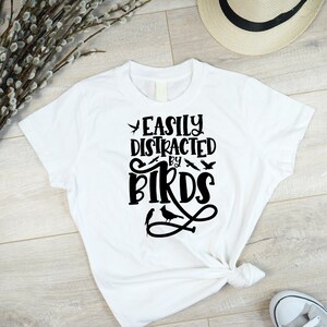 Puede incluir: Camiseta blanca con texto negro que dice "Easily Distracted by Birds" con un gr&aacute;fico de p&aacute;jaro.