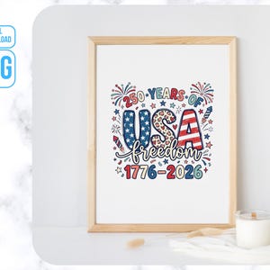 May include: A framed print with the text "250 YEARS OF USA freedom 1776-2026" in red, white, and blue. The word "USA" is in a patriotic design with stars and stripes. Fireworks and stars surround the text. A candle sits in front of the frame.