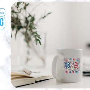 May include: A white ceramic mug with a patriotic design. The mug features the text "USA freedom 1776-2023" in red, white, and blue, with a festive fireworks design. The mug is on a white surface.