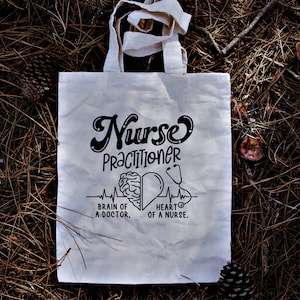 May include: White tote bag with black text that reads "Nurse Practitioner" with a brain and heart graphic and the text "Brain of a Doctor. Heart of a Nurse."