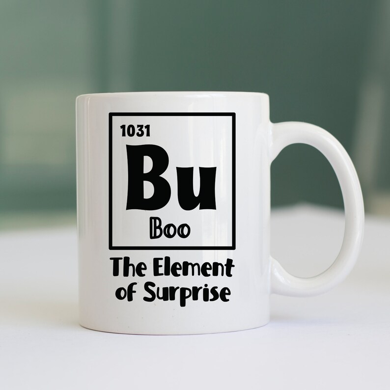 K&ouml;nnte beinhalten: Wei&szlig;e Keramik-Kaffeetasse mit schwarzem Design. Das Design ist ein Periodensystem-Elementkasten mit dem Symbol "Bu" und dem Wort "Boo" darunter. Der Text "The Element of Surprise" befindet sich unterhalb des Kastens.
