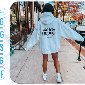 Puede incluir: Una sudadera con capucha blanca con el texto "Ponte la armadura completa de Dios" y un gr&aacute;fico de la armadura de Dios. El texto "Efesios 6:10-18" tambi&eacute;n est&aacute; incluido en la sudadera.