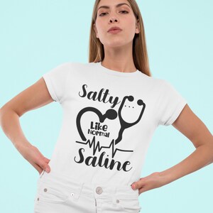 Peut inclure: T-shirt blanc avec un motif graphique noir. Le graphique repr&eacute;sente un c&oelig;ur avec un st&eacute;thoscope et le texte "Salty Like Normal Saline".