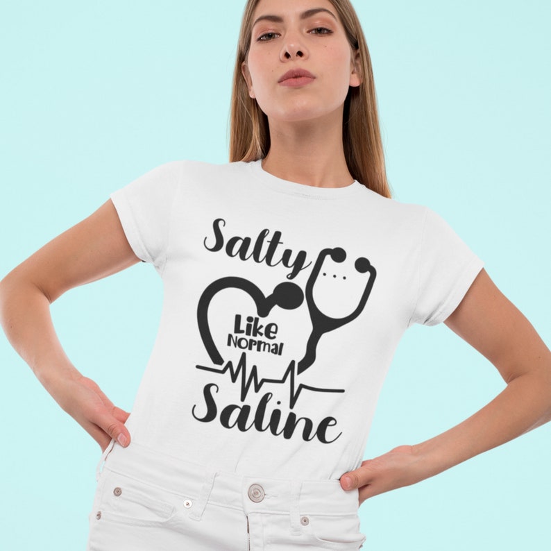 Peut inclure: T-shirt blanc avec un motif graphique noir. Le graphique repr&eacute;sente un c&oelig;ur avec un st&eacute;thoscope et le texte "Salty Like Normal Saline".