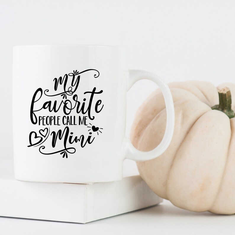 Puede incluir: Taza de cer&aacute;mica blanca con texto negro que dice "my favorite people call me Mimi" con un dise&ntilde;o de coraz&oacute;n. La taza est&aacute; sobre una superficie blanca junto a una calabaza blanca.