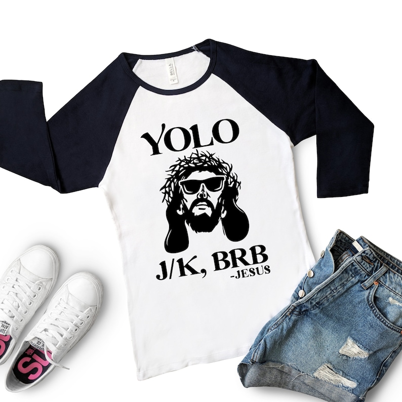 May include: A white and navy blue baseball tee shirt with a black and white graphic of Jesus wearing sunglasses and a crown of thorns. The text "YOLO J/K, BRB -JESUS" is printed on the shirt.