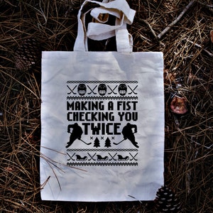 Puede incluir: Bolsa de tela blanca con texto y gr&aacute;ficos negros. El texto dice "Making a fist checking you twice". Los gr&aacute;ficos son jugadores de hockey y palos.