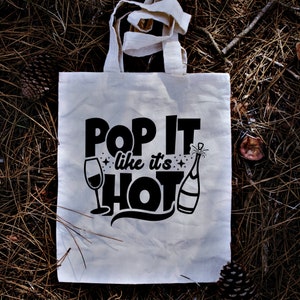 May include: A white canvas tote bag with black text that reads "Pop It Like It's Hot" with a wine glass and champagne bottle graphic.