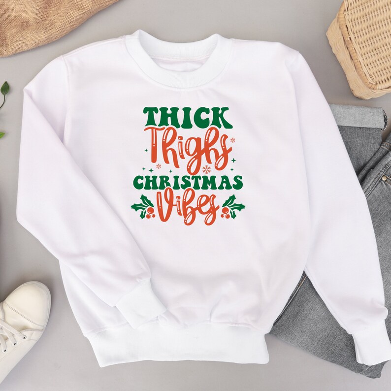 Puede incluir: Sudadera blanca con el texto "THICK THIGHS CHRISTMAS VIBES" en letras verdes y rojas con acebo y bayas.