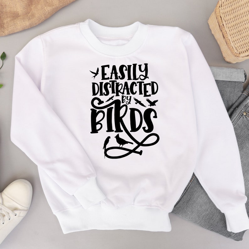 Puede incluir: Sudadera blanca con texto negro que dice "Easily Distracted by Birds". El texto est&aacute; en una fuente estilizada con peque&ntilde;as siluetas de p&aacute;jaros que lo rodean.