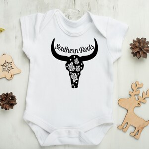 Puede incluir: Un body blanco para beb&eacute; con un dise&ntilde;o de cr&aacute;neo de toro negro con lunares blancos y rosas. El texto "Southern Roots" est&aacute; por encima del cr&aacute;neo.
