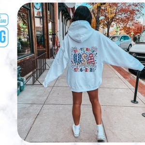 May include: A white hoodie with a colourful graphic on the back that says "USA Freedom 1776-2023". The design features red, white, and blue stars and stripes. The hoodie is worn by a person standing on a pavement.