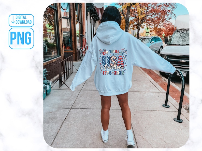 May include: A white hoodie with a colourful graphic on the back that says "USA Freedom 1776-2023". The design features red, white, and blue stars and stripes. The hoodie is worn by a person standing on a pavement.