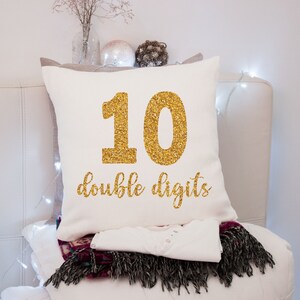 May include: White pillow with gold glitter lettering that says "10 double digits".