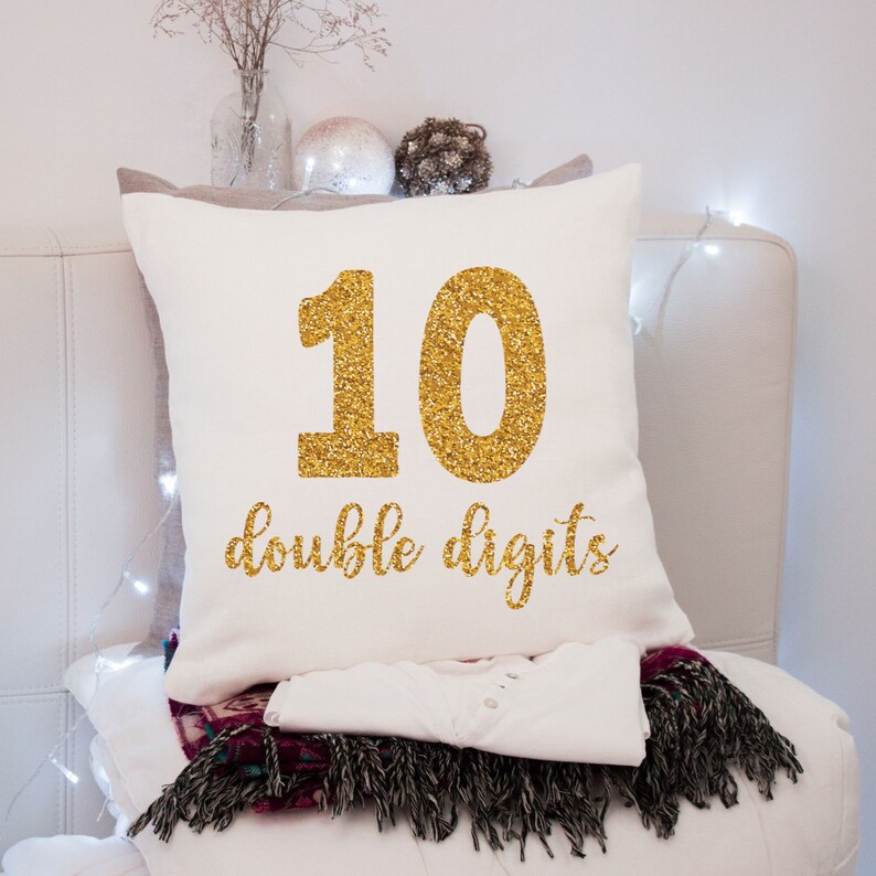 May include: White pillow with gold glitter lettering that says "10 double digits".