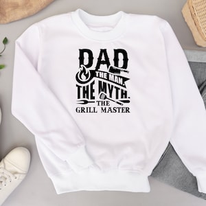 Puede incluir: Sudadera blanca con texto negro que dice "Dad The Man The Myth The Grill Master" con un gr&aacute;fico de llama y herramientas de parrilla.