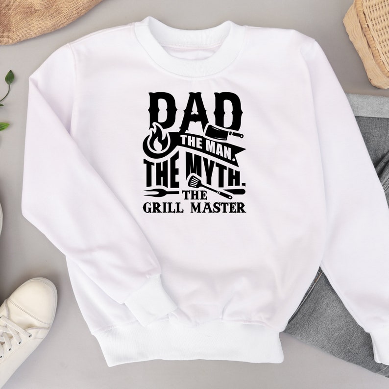 Puede incluir: Sudadera blanca con texto negro que dice "Dad The Man The Myth The Grill Master" con un gr&aacute;fico de llama y herramientas de parrilla.