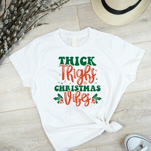 Puede incluir: Camiseta blanca con un gr&aacute;fico rojo y verde que dice "Thick Thighs Christmas Vibes".