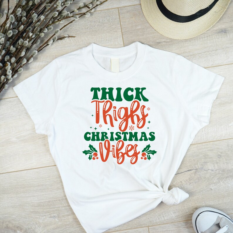 Puede incluir: Camiseta blanca con un gr&aacute;fico rojo y verde que dice "Thick Thighs Christmas Vibes".