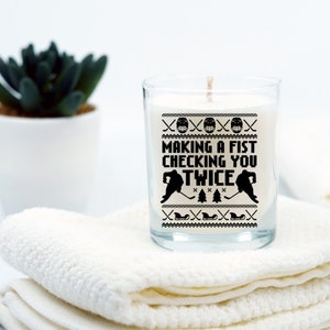 Puede incluir: Una vela blanca en un tarro de vidrio transparente con un dise&ntilde;o en blanco y negro que dice "Making a fist checking you twice". El dise&ntilde;o presenta dos jugadores de hockey y un palo de hockey.