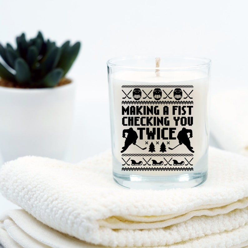 Puede incluir: Una vela blanca en un tarro de vidrio transparente con un dise&ntilde;o en blanco y negro que dice "Making a fist checking you twice". El dise&ntilde;o presenta dos jugadores de hockey y un palo de hockey.