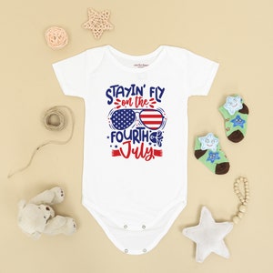 May include: White baby onesie with a red, white, and blue graphic that says "Stayin' Fly on the Fourth of July" with sunglasses that have the American flag on them.
