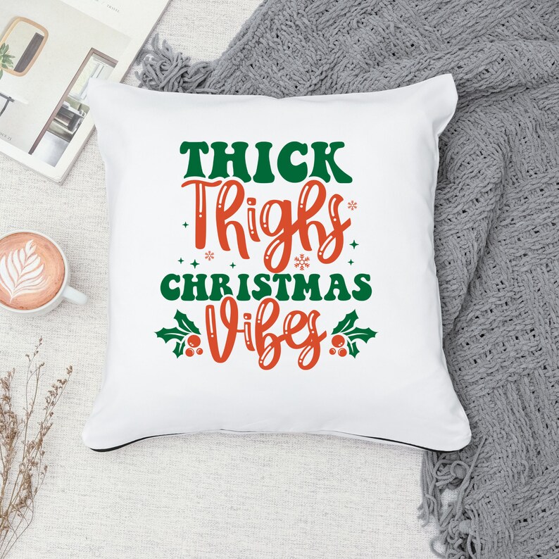 Puede incluir: Almohada blanca con el texto "THICK THIGHS CHRISTMAS VIBES" en letras rojas y verdes. El texto est&aacute; rodeado de hojas de acebo verdes y bayas rojas.