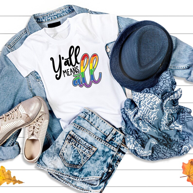 May include: White t-shirt with the text "Y'all means all" in black lettering with a rainbow arc underneath. The shirt is laid out on a white wooden surface with a denim jacket, blue jeans, a blue hat, and a blue scarf.